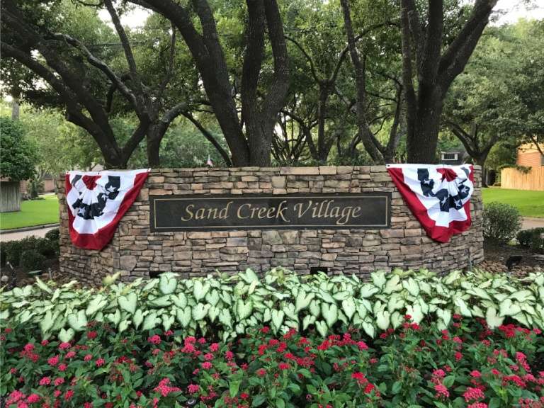 Sand Creek Village entrance sign with patriotic bunting and white seasonal flowers