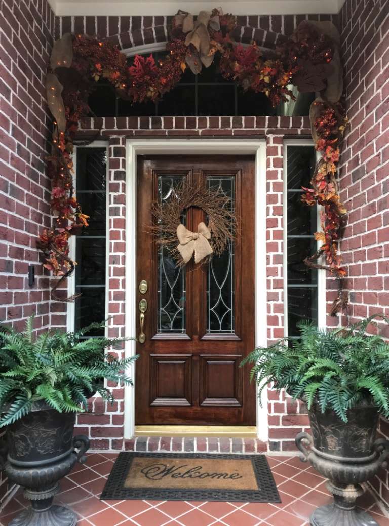 Brick front entry with autumn garland and a burlap bow wreath flanked by potted ferns